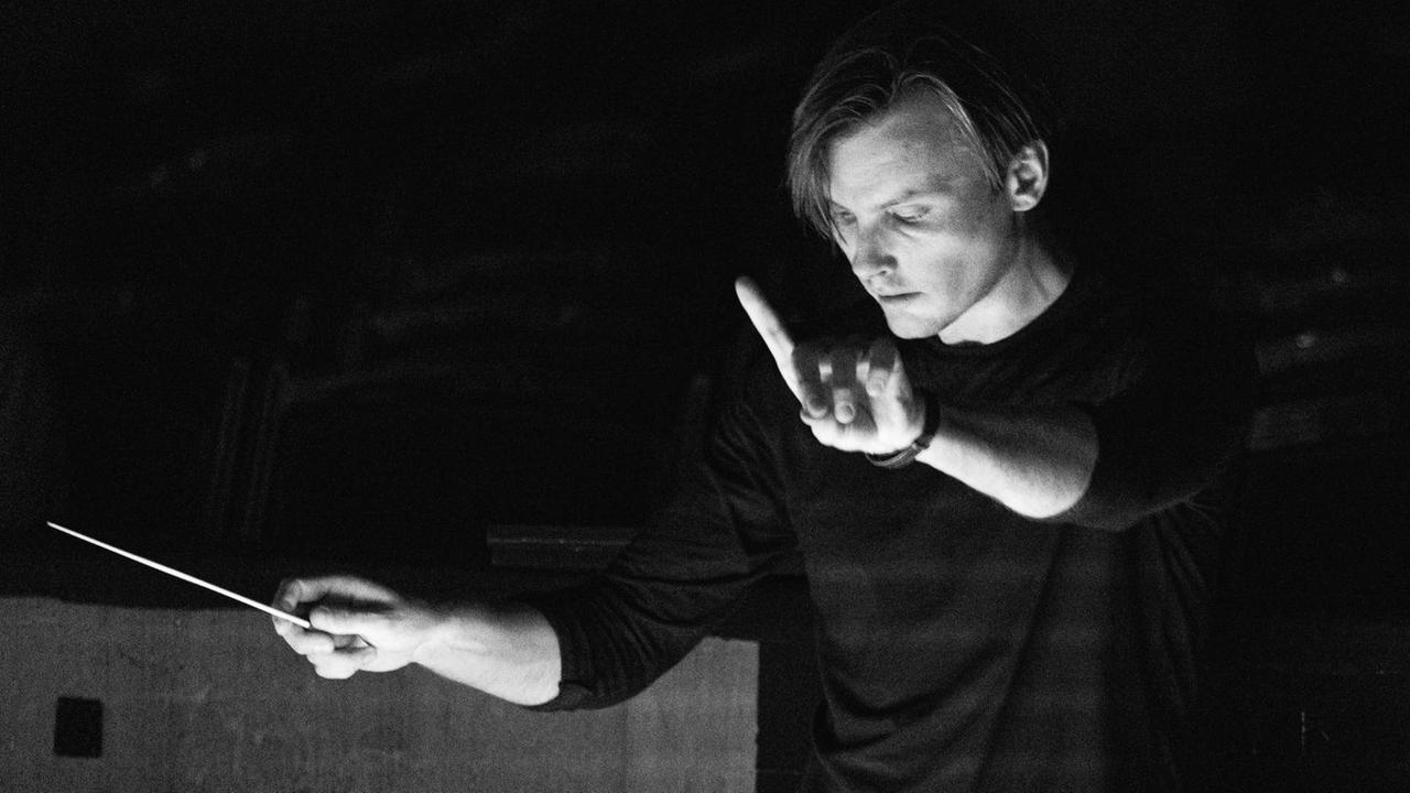 Auf der Schwarz-weiß-Fotografie hebt der belarussische Dirigent die rechte Hand mit dem Taktstock, während er konzentriert den Blick gesenkt hat und mit dem linken Zeigefinger ein Zeichen gibt.