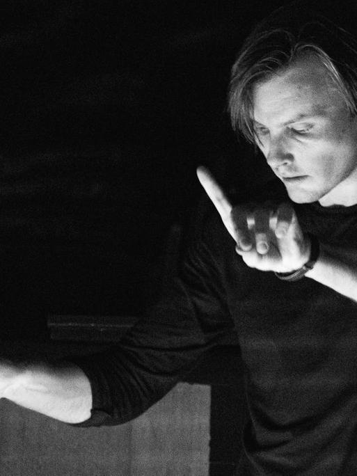 Auf der Schwarz-weiß-Fotografie hebt der belarussische Dirigent die rechte Hand mit dem Taktstock, während er konzentriert den Blick gesenkt hat und mit dem linken Zeigefinger ein Zeichen gibt.