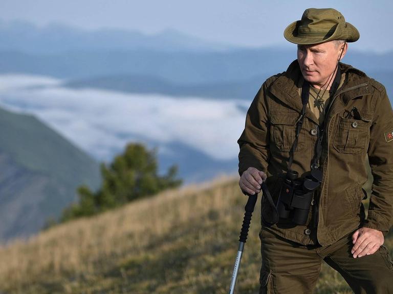 Putin mit Wanderstock und Fernglas in Outdoor-Bekleidung. Im Hintergrund öffnet sich der Blick von oben in eine Tagebene hinein.