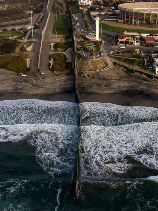 Luftansicht des Grenzzauns zwischen den USA und Mexiko, der durch den Strand bis mitten hinein ins Wasser reicht.
