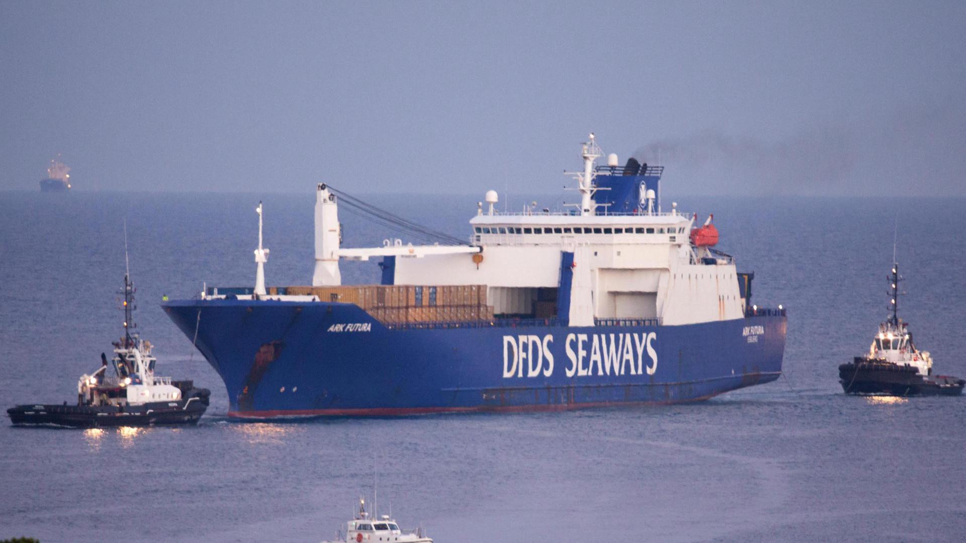 Ein blaues großes Schiff liegt vor einem Hafen, flankiert von zwei kleineren Schlepperschiffen. Ein blaues großes Schiff liegt vor einem Hafen, flankiert von zwei kleineren Schlepperschiffen.
