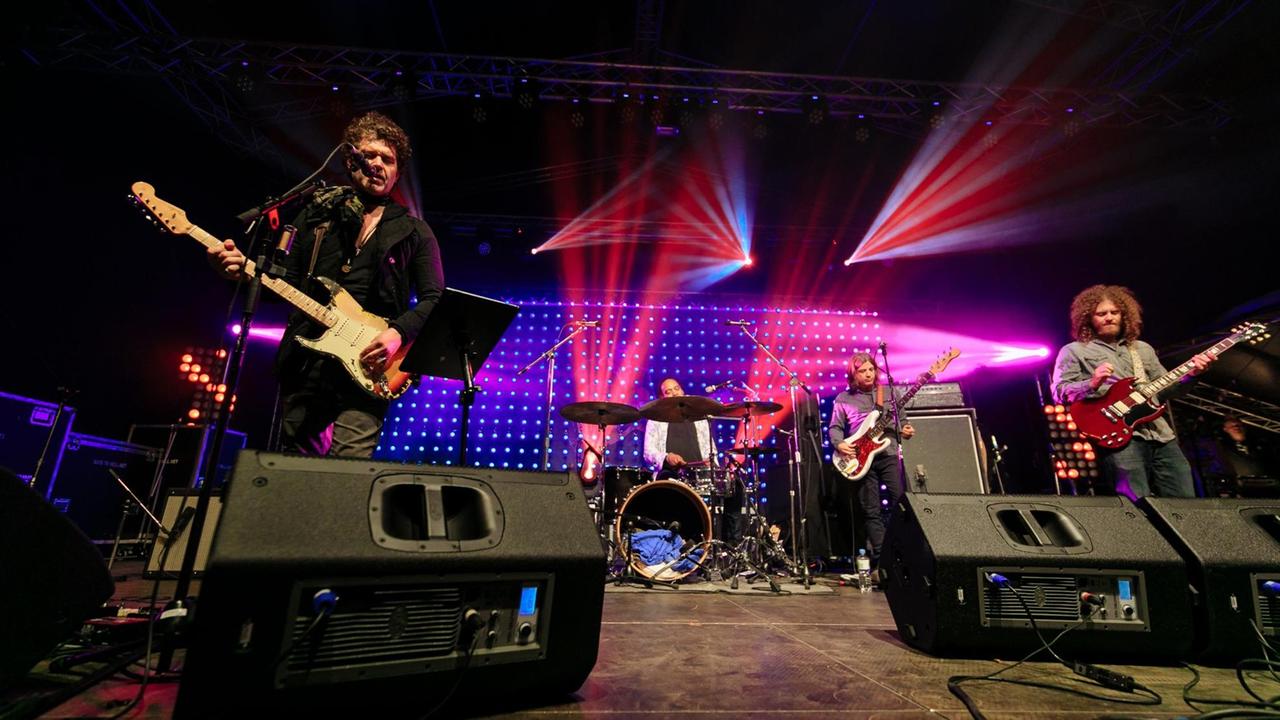 Eine Band steht auf einer bunt beleuchteten Bühne und spielt, am vorderen Bühnenrand schwarze Monitorboxen