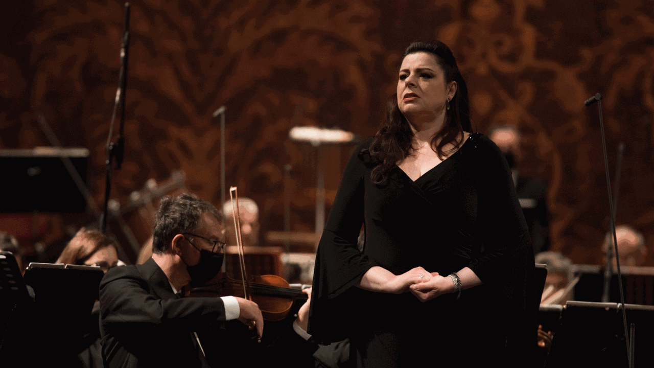 Daniela Barcellon steht in dunklem Abendkleid vor dem Orchester, das weitesgehend Mundschutz trägt.