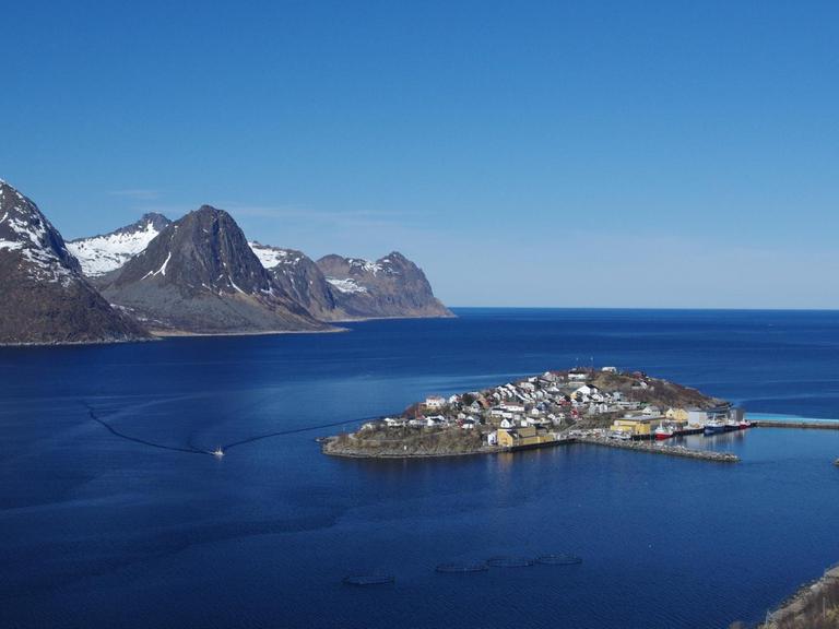 Ein auf einer Insel gelegenes Fischerdorf in Norwegen. Im Hintergrund sind Berge und der Horizont zu sehen.