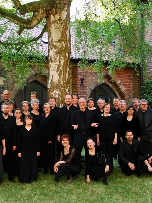 40 Chormitglieder in schwarzer Konzertkleidung haben sich unter einer großen Birke auf der Wiese im Innenhof des Ratzeburger Doms versammelt.