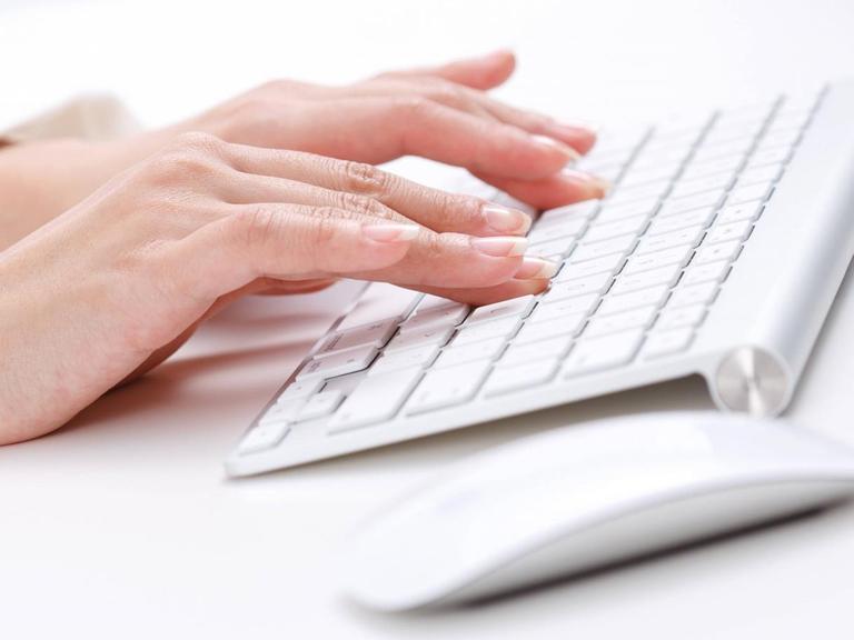 Frauenhände tippen auf einer weißen Computertastatur. Rechts daneben liegt eine weiße Computermaus.