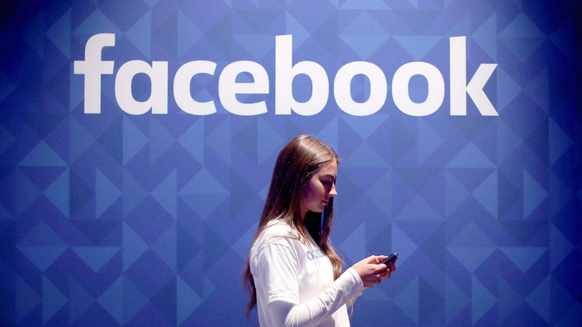 Eine Frau schaut auf ihr Smartphone, während sie an einer blauen Wand mit Facebook-Logo vorbei läuft.