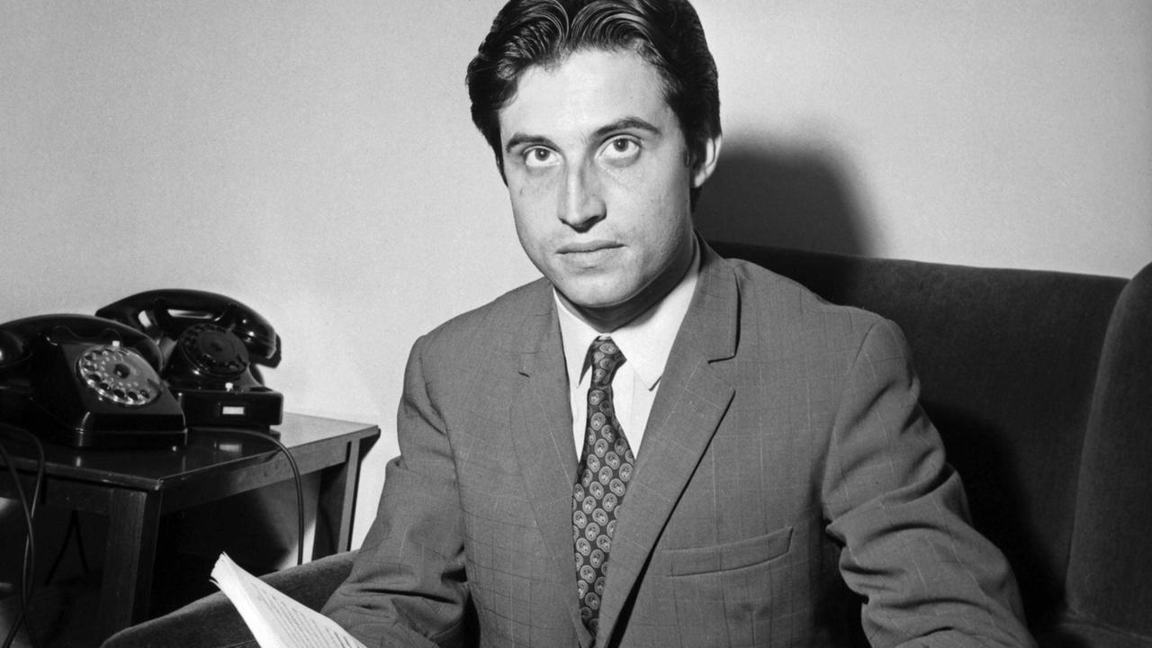 Riccardo Muti sitzt als junger Mann 1967 auf einem Sofa und hält ein Buch in der Hand, während er spontan zur Kamera schaut.