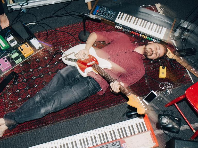 Ein Mann liegt, umringt von Instrumenten, mit seiner E-Gitarre auf dem Rücken auf einem Teppich.