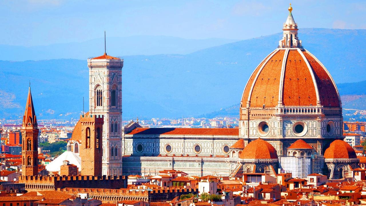 Ein Farbfoto zeigt das Panorama von Florenz mit mit rotziegelten Dächern. Die Hügel der Toskana im Hintergrund Davor die Kuppel des Doms un dem Campanile