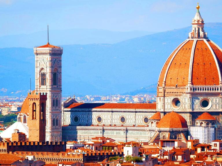 Ein Farbfoto zeigt das Panorama von Florenz mit mit rotziegelten Dächern. Die Hügel der Toskana im Hintergrund Davor die Kuppel des Doms und der Campanile
