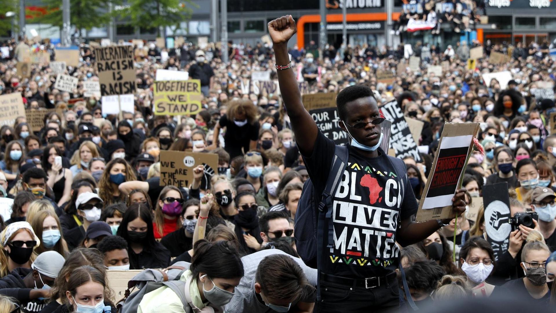 Vor einer großen sitzenden Menschenmenge mit Gesichtsmasken steht ein Schwarzer mit erhobener Faust. Auf seinem T-Shirt steht in bunten Buchstaben "Black Lives Matter".