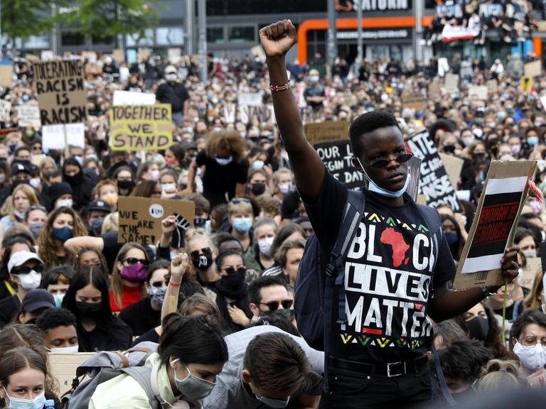 Vor einer großen sitzenden Menschenmenge mit Gesichtsmasken steht ein Schwarzer mit erhobener Faust. Auf seinem T-Shirt steht in bunten Buchstaben "Black Lives Matter".