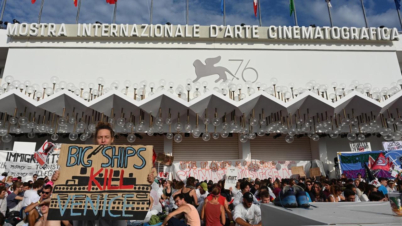 Der Palazzo del Cinema in Venedig mit dem Logo der 76. Filmfestspiele in Venedig. Protestierende sitzen auf dem roten Teppich und halten Plakate in die Höhe, eines davon mit der Aufschrift: "Big Ships Kill Venice" 