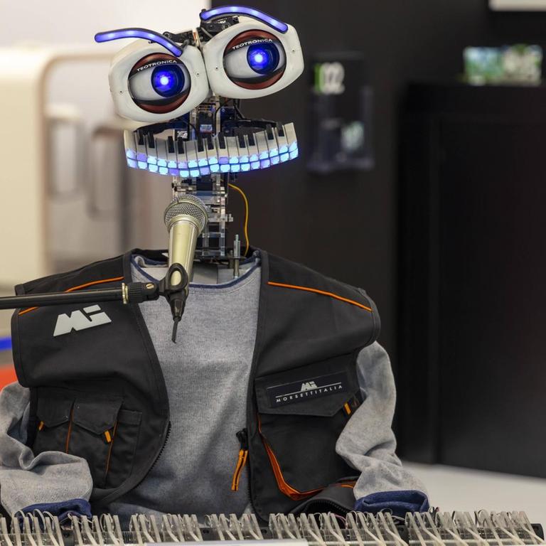 Ein Roboter mit Tastaturmund und großen Augen sitzt vor einer Klaviatur und Mikrofon und scheint zu musizieren. 