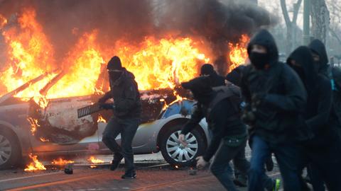 Demonstranten mit schwarzen Kapuzen und Schals, im Hintergrund brennt ein Polizei-Auto