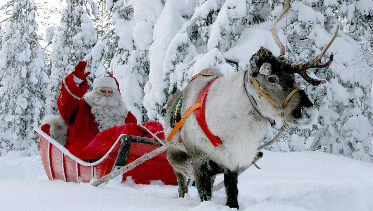 Ein als Weihnachtsmann oder Nikolaus verkleideter Mann sitzt auf einem Schlitten, der von einem Rentier gezogen wird. Im Hintergrund sieht man schneebedeckte Tannen.