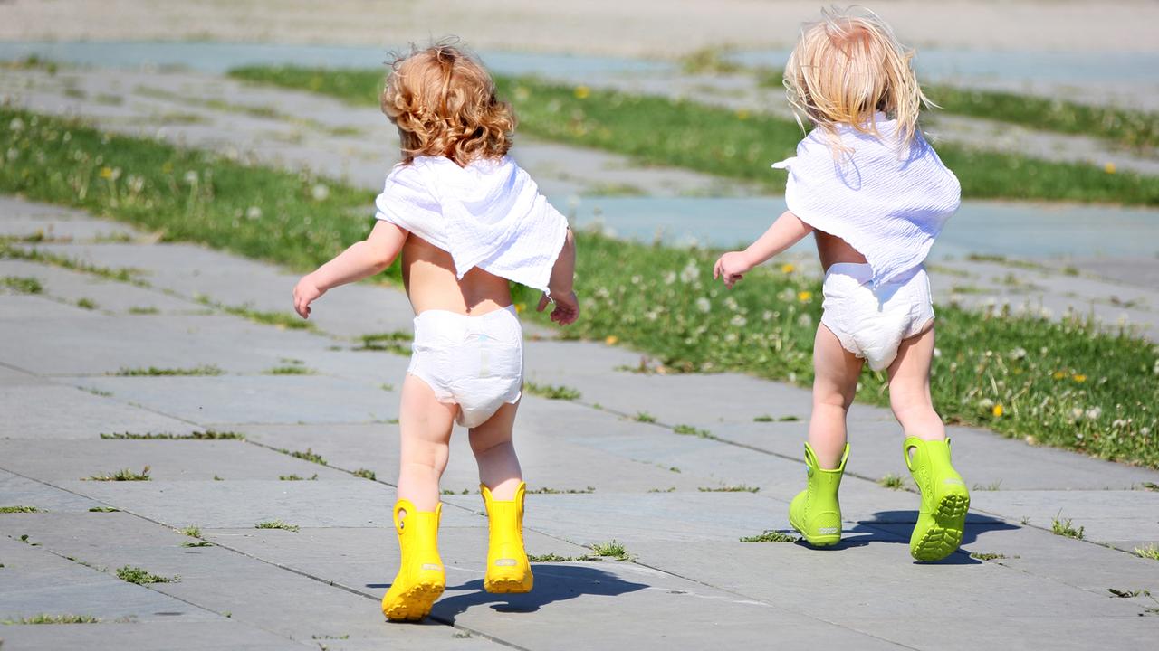 In Windeln und Gummistiefeln laufen zwei kleine Mädchen bei frühsommerlichen Temperaturen über einen Platz.