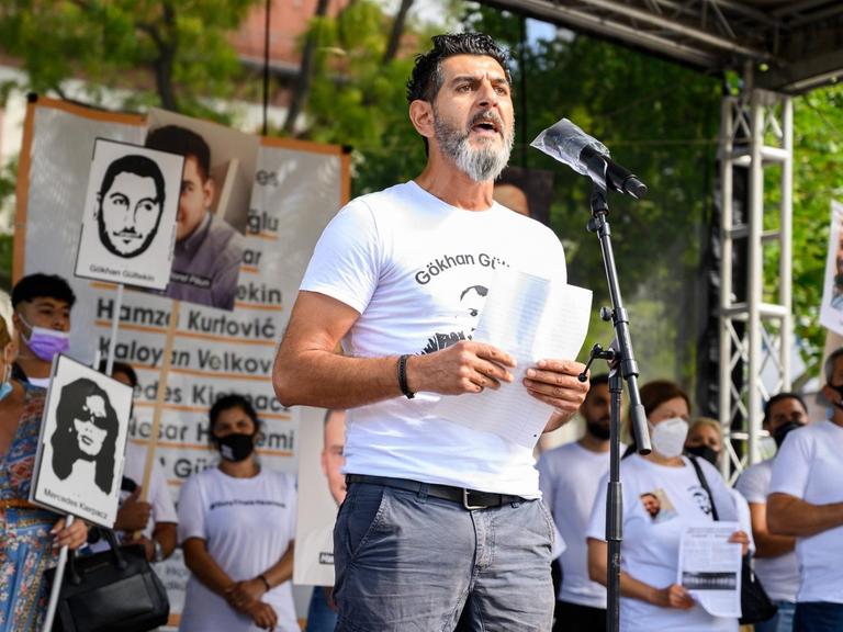 Cetin Gültekin spricht auf einer Freiluftbühne im Sonnenschein in ein Mikrofon. Auf seinen T-Shirt steht der Name seines ermordeten Bruders Gökhan Gültekin sowie eine Zeichnung dessen Gesichts.