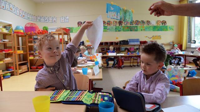 Ein Kind mit Down-Syndrom zeigt stolz ein selbst gemaltes Bild. Daneben sitzt ein Kind ohne Behinderung.