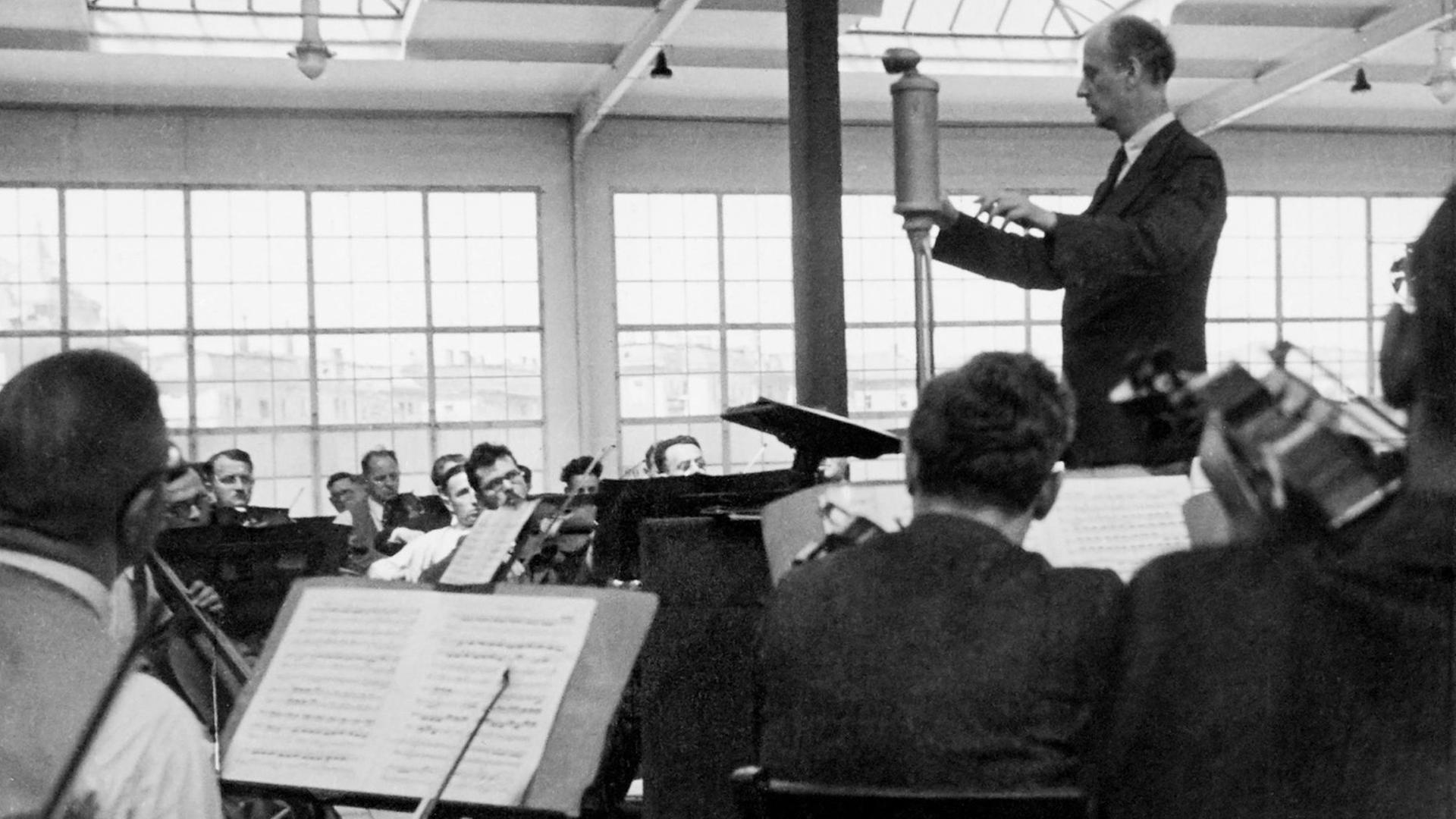Schwarz-Weiß-Fotografie eines Menschen im Frack, der ein Orchester dirigiert. In der Mitte des Raums steht ein Mikrofon.