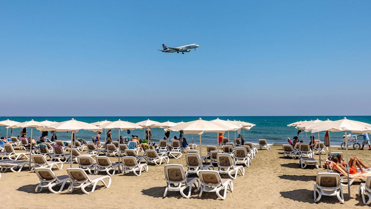 Ein Flugzeug fliegt bei strahlendem Sonnenschein tief über einem Strand mit Reihen voller Liegen und Touristen.