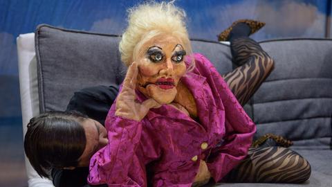 Eine lebensgroße Puppe in einer roten Bluse, aus dem Stück Volksvernichtung , liegt auf einem Sofa und wird von einer Schauspielerin bespielt.