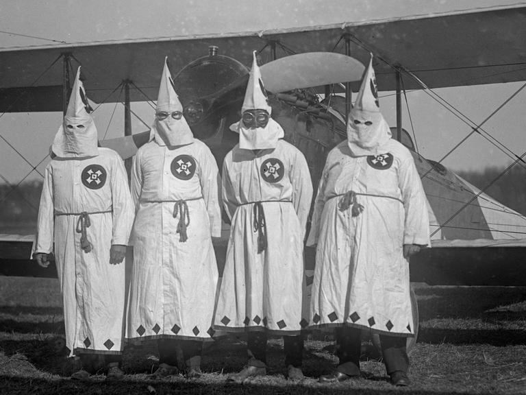 Vier Männer in weißen Kutten mit spitzzulaufenden Hüten stehen vor einer alten Propellermaschine. Das Bild ist schwarz-weiß.