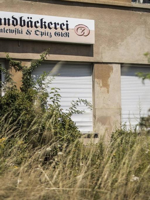 Eine aufgegebene Bäckerei in Klitten. Das Schild des Geschäfts ist noch zu sehen, aber die Rolläden sind herabgelassen und der Zugang zum Gebäude ist von Unkraut überwuchert.