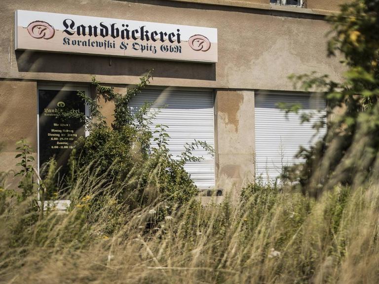 Eine aufgegebene Bäckerei in Klitten. Das Schild des Geschäfts ist noch zu sehen, aber die Rolläden sind herabgelassen und der Zugang zum Gebäude ist von Unkraut überwuchert.