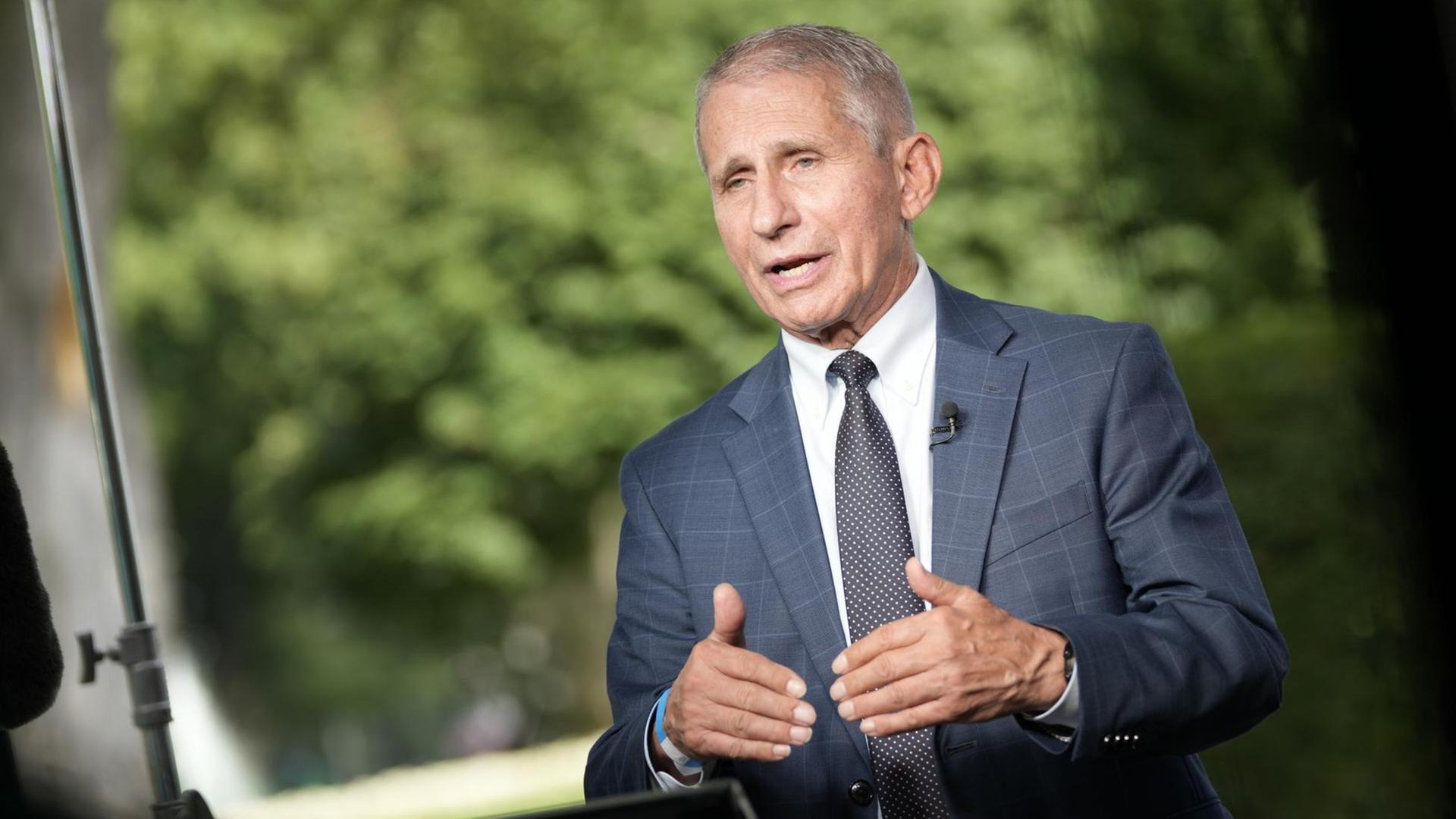 Zu sehen ist der Immunologe Anthony Fauci. Er trägt einen blauen Anzug, ein weißes Hemd und eine schwarze Krawatte. Er steht an einem Rednerpult und gestikuliert mit seinen Händen.