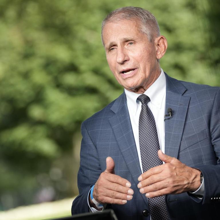 Zu sehen ist der Immunologe Anthony Fauci. Er trägt einen blauen Anzug, ein weißes Hemd und eine schwarze Krawatte. Er steht an einem Rednerpult und gestikuliert mit seinen Händen.