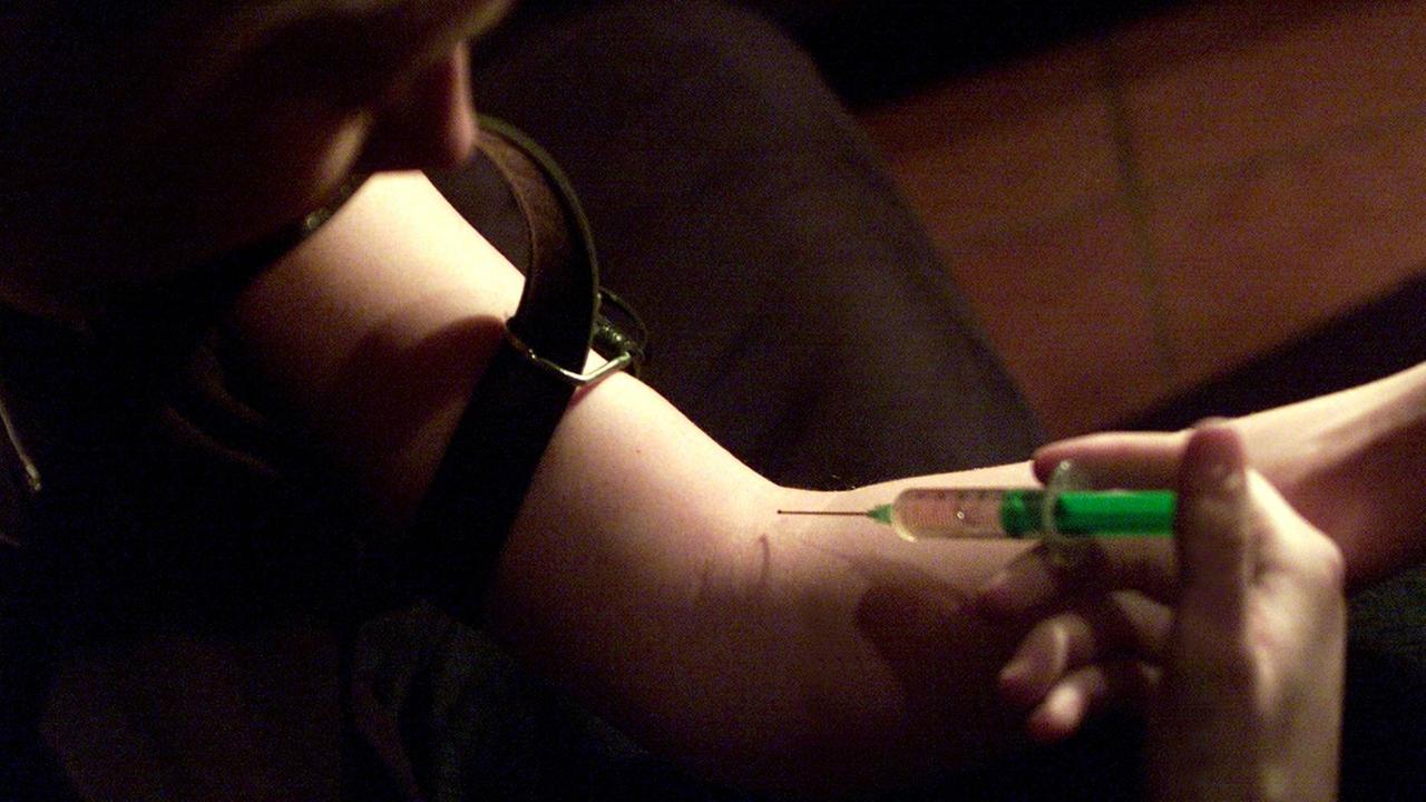 Eine Drogenabhängige setzt sich einen Schuss: Der Arm ist mit einem Lederriemen abgebunden, mit der anderen Hand sticht sie mit einer grünen Spritze in die Armbeuge.