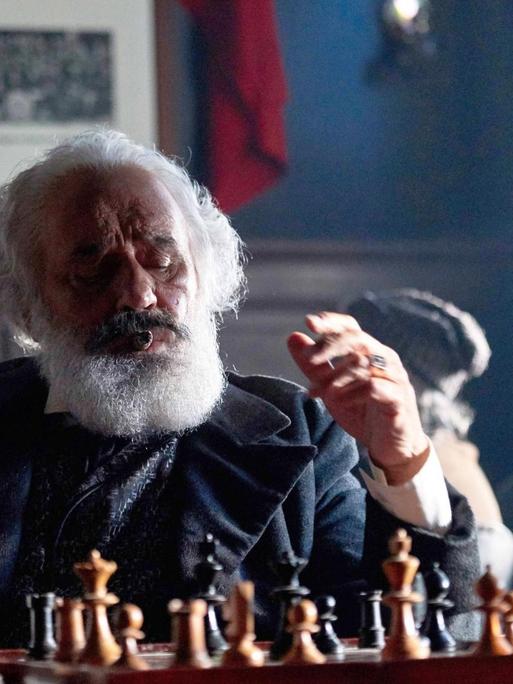 Mario Adorf, in der Rolle des Karl Marx, sitzt mit einem Schachbertt und einem Glas Alkohol am Tisch.