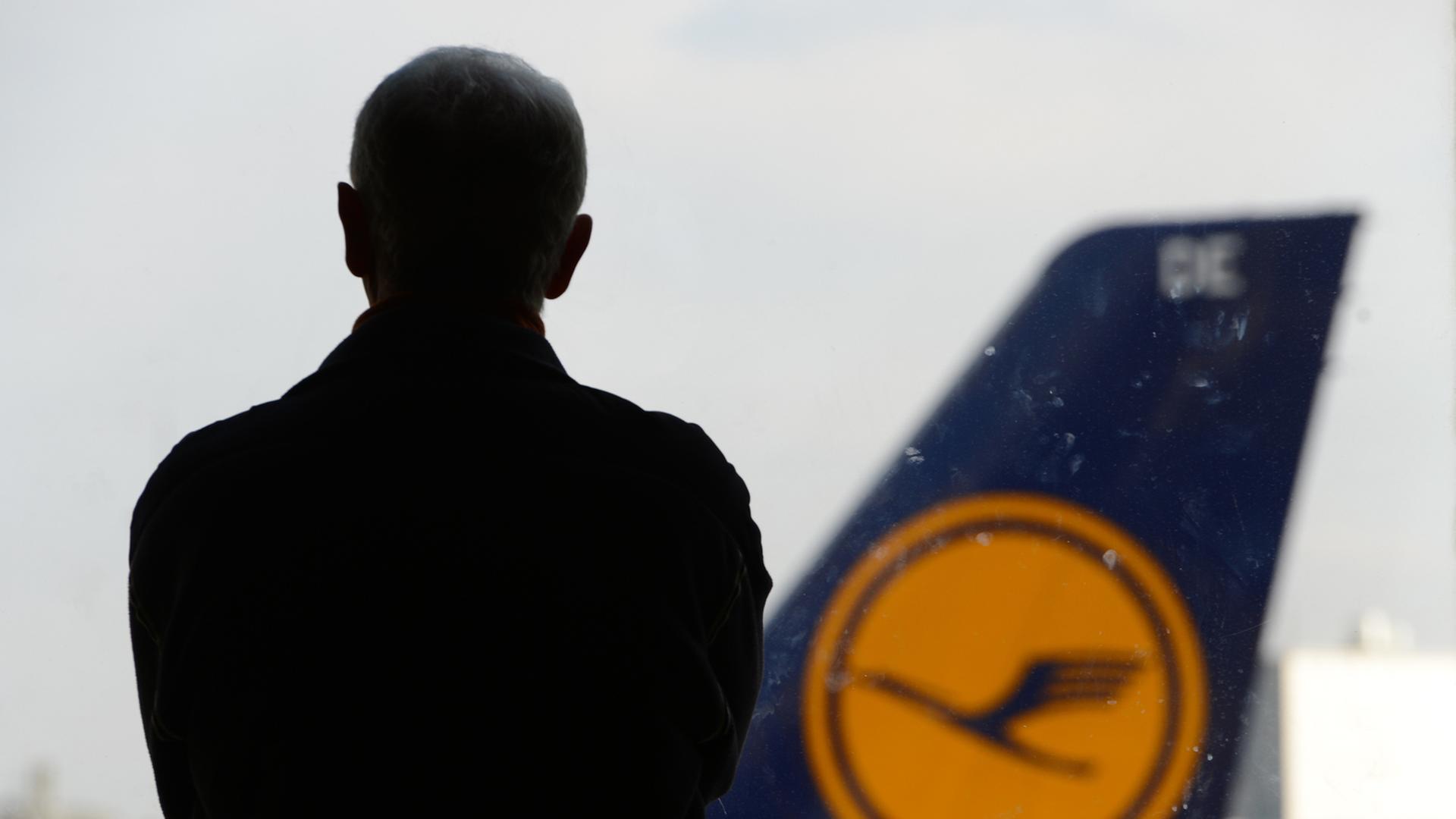 Ein Mann wendet der Kamera den Rücken zu und blickt auf eine Flugzeugheck, auf dem das Logo der Lufthansa zu sehen ist.