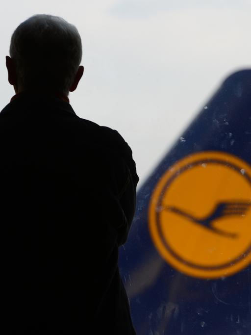 Ein Mann wendet der Kamera den Rücken zu und blickt auf eine Flugzeugheck, auf dem das Logo der Lufthansa zu sehen ist.