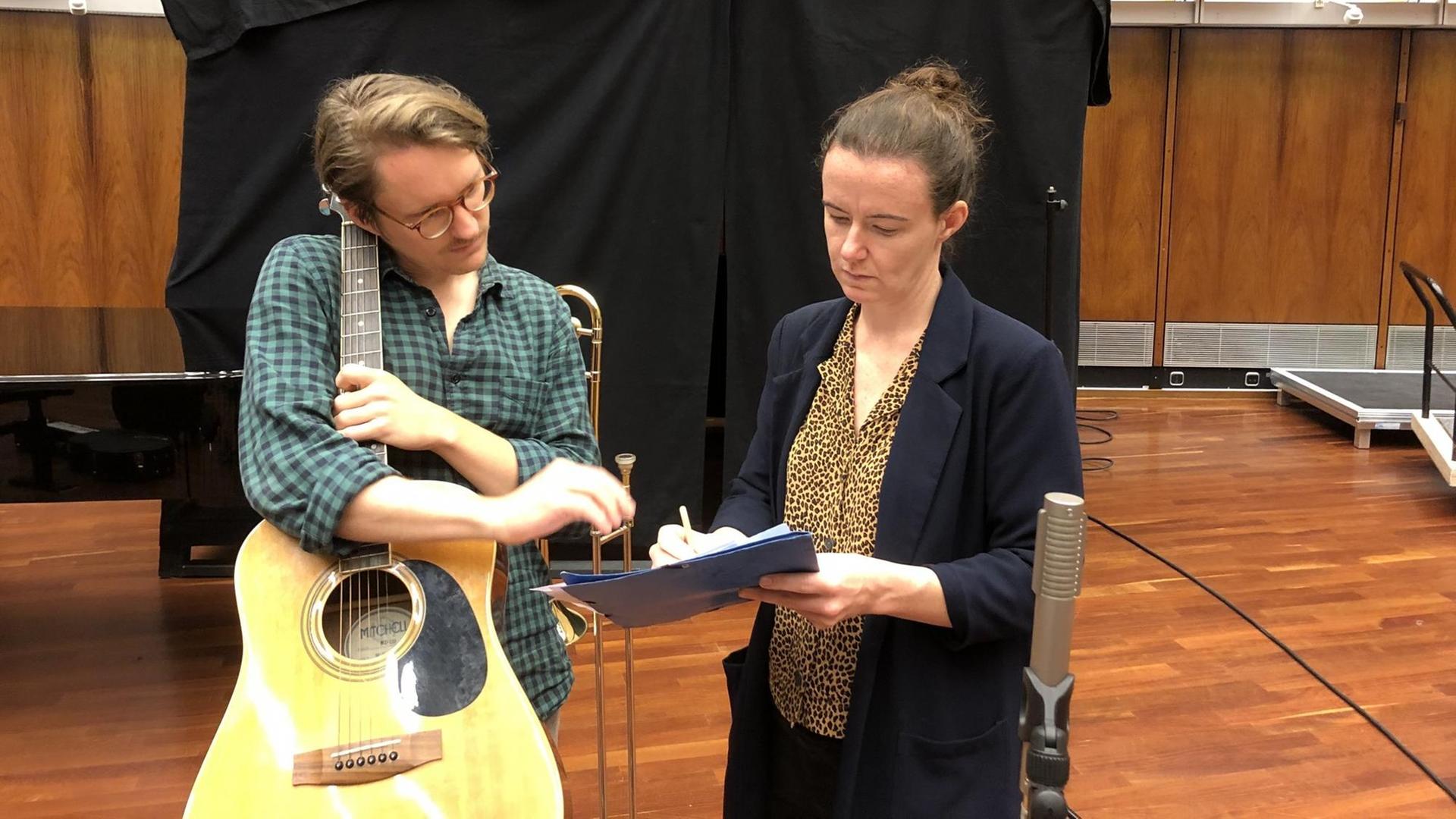 Ein Mann mit Gitarre im Arm schaut auf ein Papier, das eine Frau neben ihm in der Hand hält