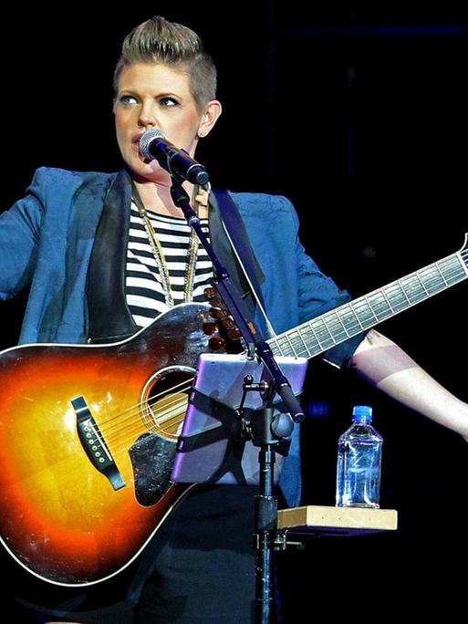 Eine Frau mit Gitarre, gestreiftem Shirt und blauem Blazer steh mit ausgebreiteten Armen am Mikrofon auf einer Bühne.