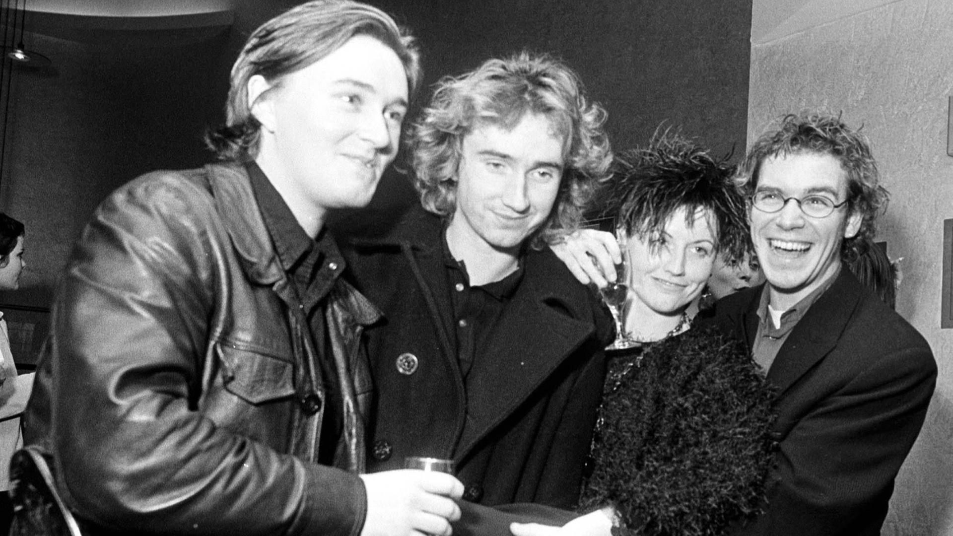 Die irische Rockgruppe The Cranberries im Jahr 1997. Die vier Bandmitglieder inklusive der inzwischen verstorbenen Dolores O'Riordan stehen nebeneinander und lächeln, manche haben ein Sektglas in der Hand. Die irische Rockgruppe The Cranberries im Jahr 1997. Die vier Bandmitglieder inklusive der inzwischen verstorbenen Dolores O'Riordan stehen nebeneinander und lächeln, manche haben ein Sektglas in der Hand.