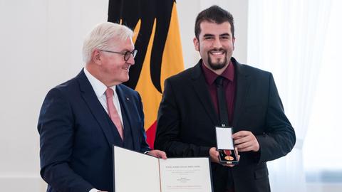 Burak Yilmaz präsentiert freudig den Verdienstorden der Bundesrepublik Deutschland, den er von Bundespräsident Frank-Walter Steinmeier verliehen bekommt. Steinmeyer hält, ebenfalls lächelnd, die Urkunde in die Kamera. Steinmeier würdigt Ehrenamtliche für herausragendes Engagement für die Gedenk- und Erinnerungskultur in Deutschland.