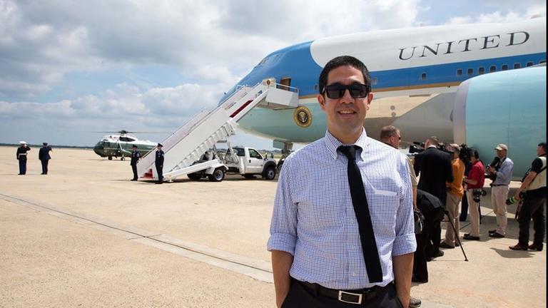 Ein junger Mann mit Sonnenbrille, kurzärmligem Hemd und schwarzer Krawatte steht am Flughafen vor der Maschine des US-Präsidenten.