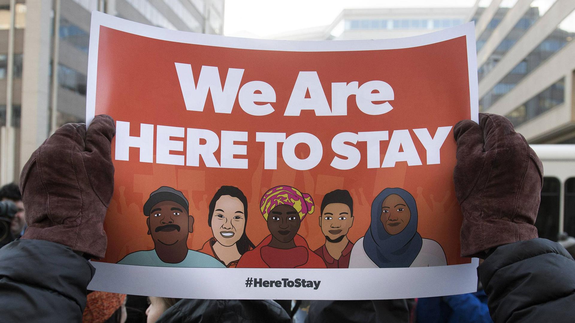 Ein Demonstrant hält ein Plakat mit dem Spruch "We are here to stay" hoch.
