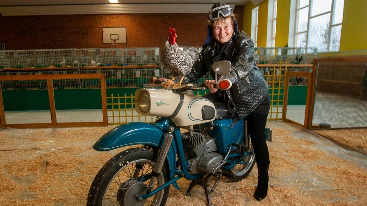 Eine Seniorin sitzt in einem Hühnerstall auf einem alten Motorrad und lächelt. Auf dem Tank des Mororrads sitzt ein Hahn.