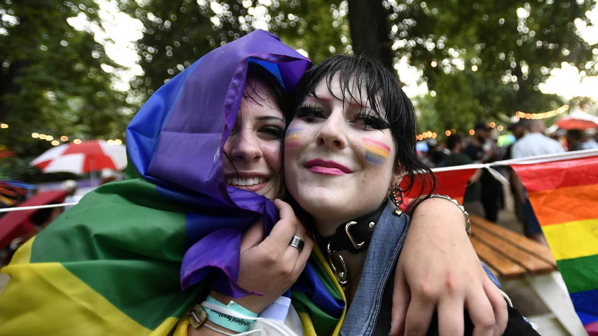 Zwei in eine Regenbogenfahne gehüllte junge Frauen umarmen sich und lächeln in die Kamera.