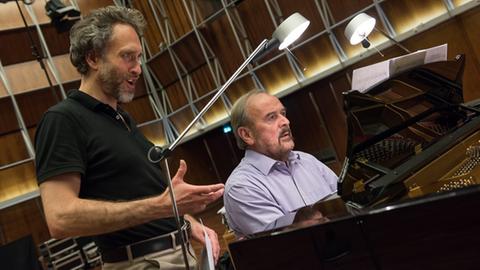 In einem mit Holz ausgekleidetem Saal steht links ein Mann mit Vollbart und schwarzem Polohemd und singt, neben ihm sitzt ein weiterer Mann an einem Flügel und spielt darauf. Beide schauen in die Noten auf dem Flügel.