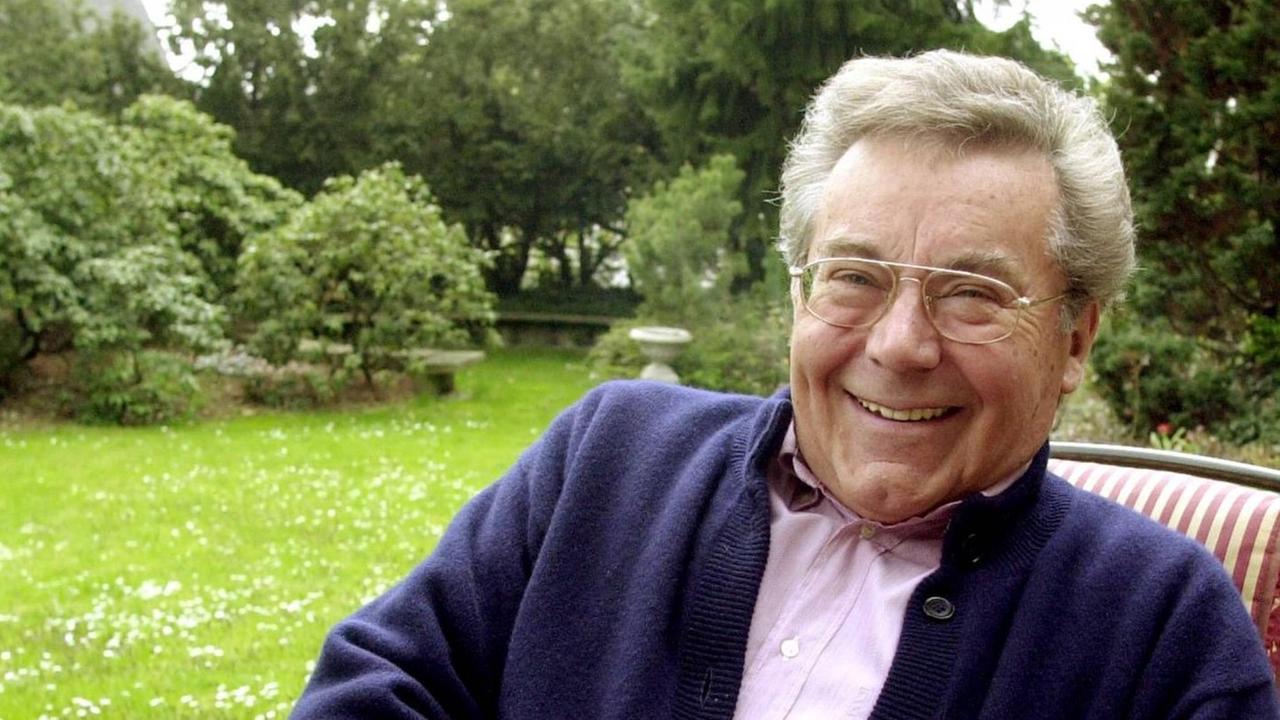 Ein ältere Herr mit Brille sitzt in einem Gartenstuhl und lacht fröhlich in die Kamera.
