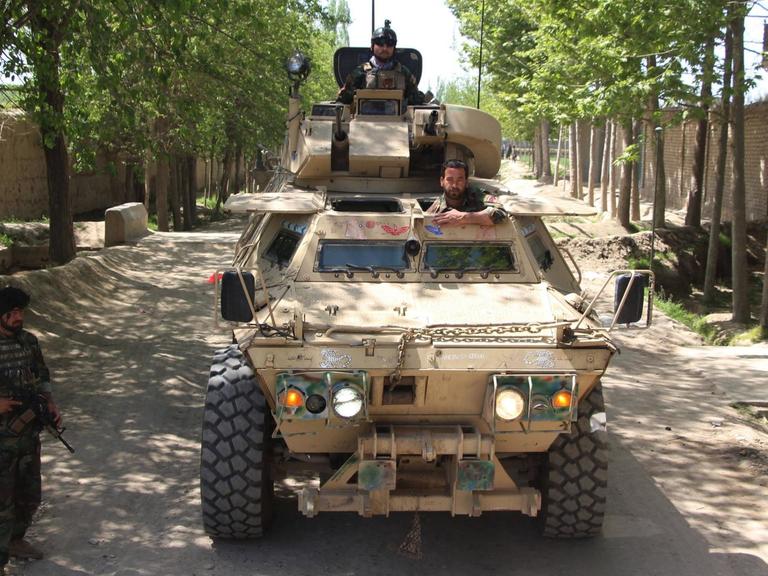 Zwei Männer schauen aus einem Panzer, der auf einer Straße steht. Daneben steht ein weiterer, bewaffneter Mann.