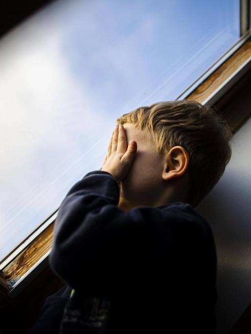 Ein kleiner Junge steht am Fenster und hält sich die Hände vor sein Gesicht.