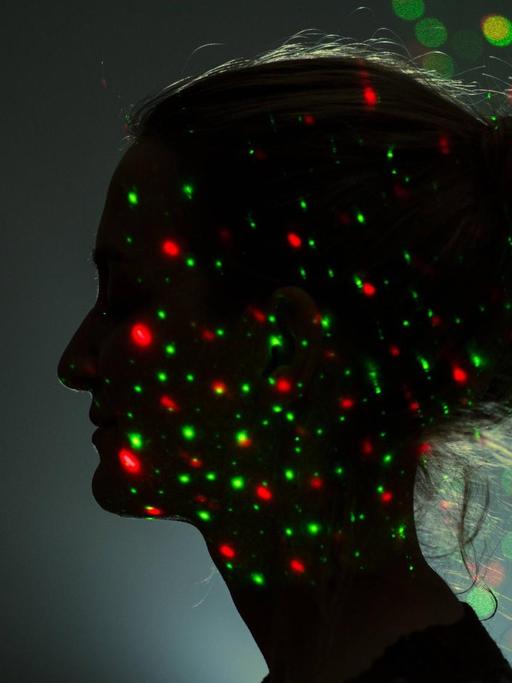 Das Profil eines Frauengesichtes ist in der Dunkelheit zu erkennen. Grüne und rote Lichjtpunkte leuchten auf ihrem Gesicht und um ihrem Kopf