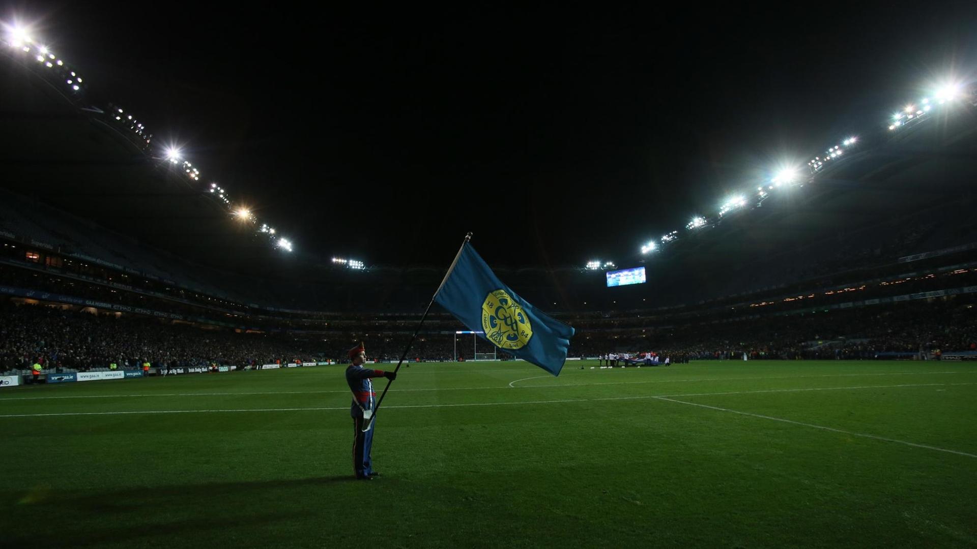 Eine Foto zeigt einen Mann in Uniform eine Flagge haltend auf leerem Rasen im mit Flutlicht beleuchteten Croke-Park-Stadion in Dublin.